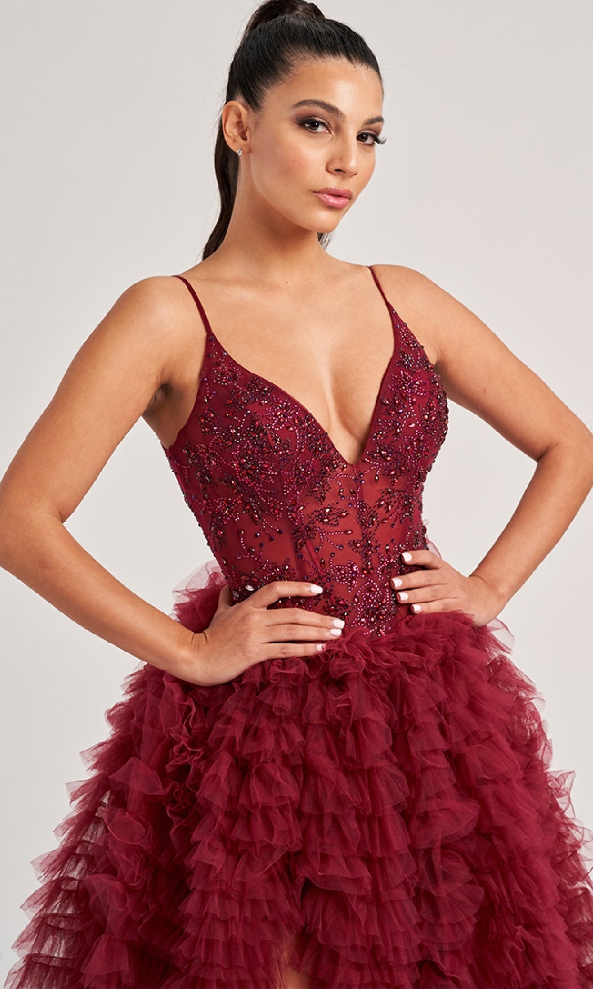 A woman poses with her hands on her hips, wearing the Long Prom Dress: Colette CL8330—a sleeveless, deep red gown featuring a fitted beaded bodice and a voluminous tiered ruffled skirt—against a plain background.